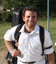 a white man with brown hair and a red beard. he is wearing white pants and a white shirt with black suspenders, looking at the camera and smiling